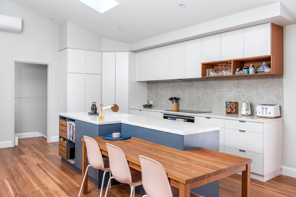 Modern inredning av ett vit vitt kök, med en undermonterad diskho, släta luckor, vita skåp, grått stänkskydd, stänkskydd i cementkakel, integrerade vitvaror, mellanmörkt trägolv, en köksö och brunt golv