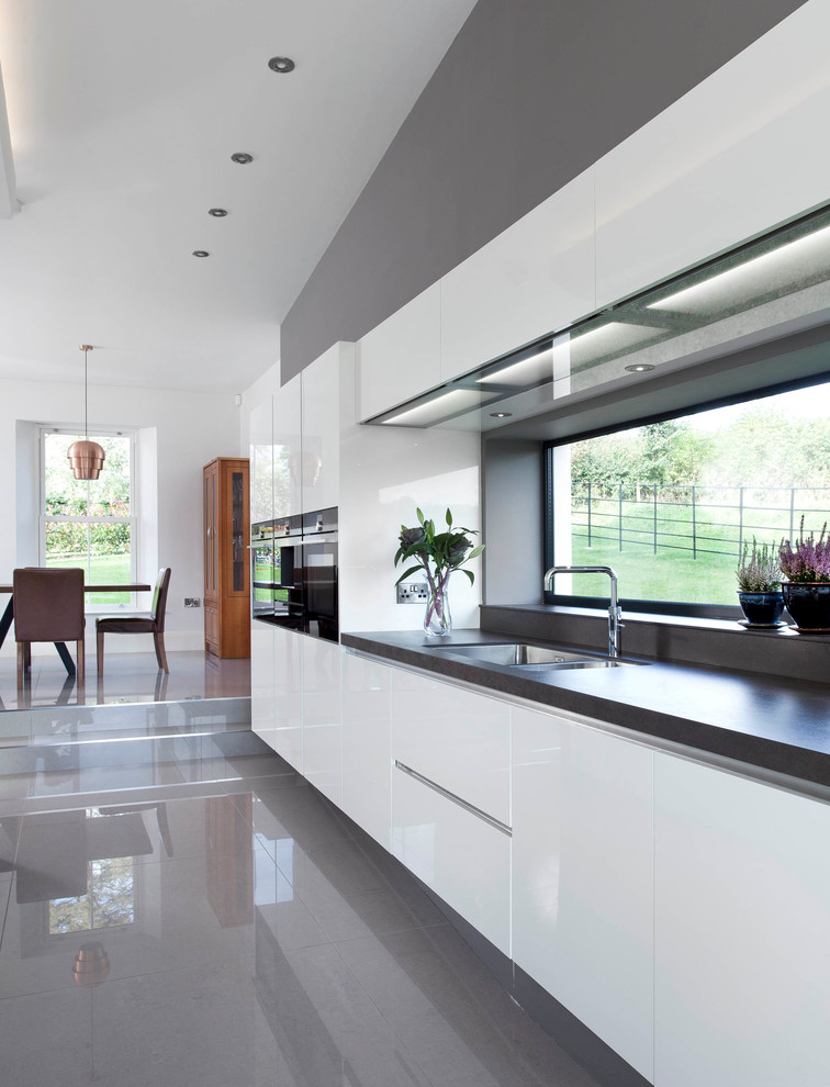 Photo of a large contemporary single-wall open plan kitchen in Belfast with an integrated sink, flat-panel cabinets, white cabinets, engineered stone countertops, grey splashback, stone slab splashback, stainless steel appliances, porcelain flooring and an island.