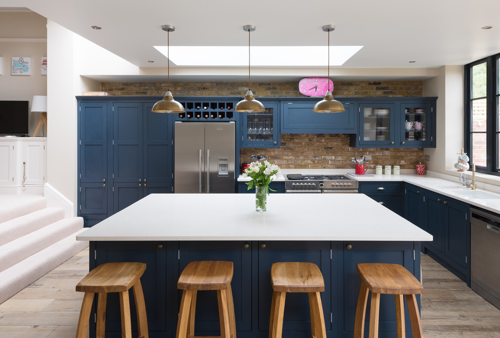 Klassische Küche in L-Form mit Schrankfronten im Shaker-Stil, blauen Schränken, hellem Holzboden, Kücheninsel, Unterbauwaschbecken, Küchenrückwand in Braun und Küchengeräten aus Edelstahl in Gloucestershire