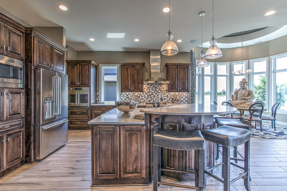 This is an example of a large traditional l-shaped kitchen/diner in Omaha with raised-panel cabinets, dark wood cabinets, composite countertops, mosaic tiled splashback, stainless steel appliances, light hardwood flooring, an island, a double-bowl sink and multi-coloured splashback.
