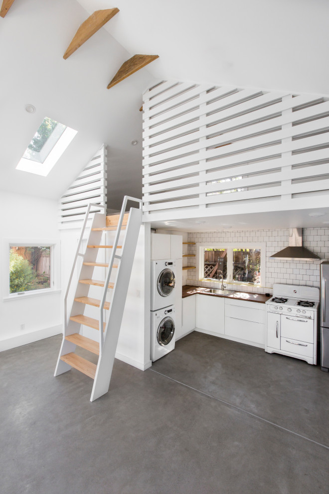 Photo of a small rural l-shaped open plan kitchen in San Francisco with a single-bowl sink, flat-panel cabinets, white cabinets, laminate countertops, white splashback, ceramic splashback, white appliances, concrete flooring, no island, grey floors, brown worktops and a vaulted ceiling.