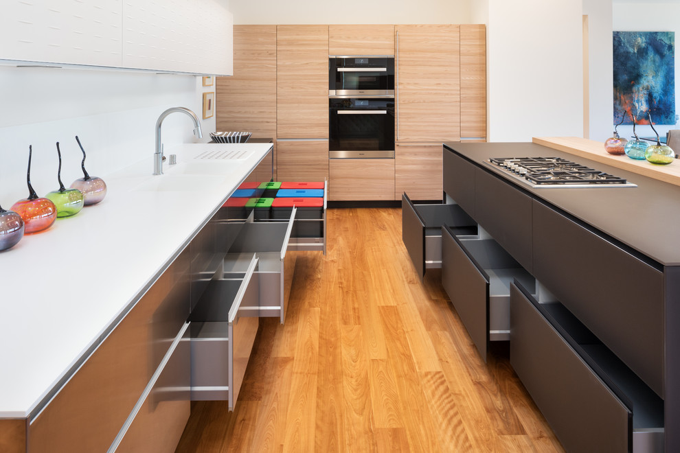 Design ideas for a large contemporary l-shaped open plan kitchen in Minneapolis with an integrated sink, flat-panel cabinets, light wood cabinets, glass worktops, white splashback, glass sheet splashback, black appliances, medium hardwood flooring and an island.
