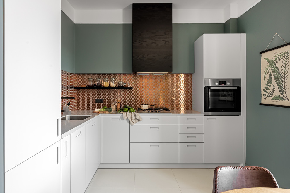 Modern inredning av ett l-kök, med en enkel diskho, släta luckor, vita skåp, stänkskydd med metallisk yta och svarta vitvaror