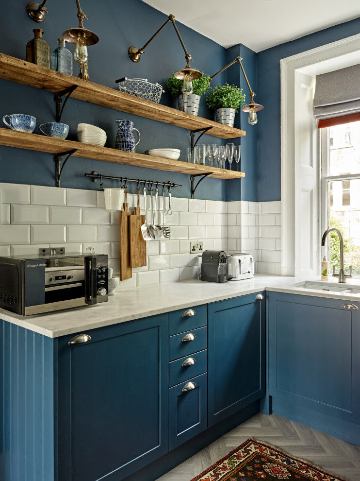 На фото: кухня в стиле неоклассика (современная классика) с врезной мойкой, фасадами в стиле шейкер, синими фасадами, белым фартуком, фартуком из плитки кабанчик, серым полом и белой столешницей