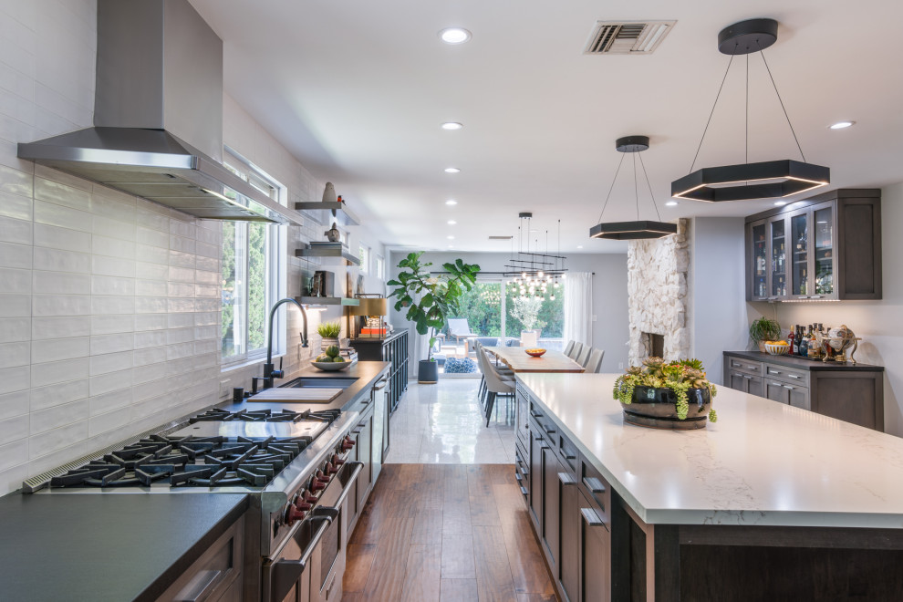 This is an example of a large contemporary kitchen/diner in Los Angeles with a submerged sink, recessed-panel cabinets, brown cabinets, composite countertops, white splashback, metro tiled splashback, stainless steel appliances, medium hardwood flooring, an island, brown floors and white worktops.