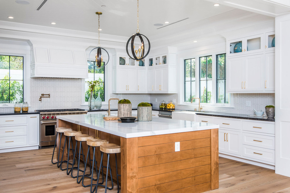 Photo of a beach style kitchen in Los Angeles with a belfast sink, shaker cabinets, white cabinets, white splashback, stainless steel appliances, medium hardwood flooring, an island, brown floors and black worktops.