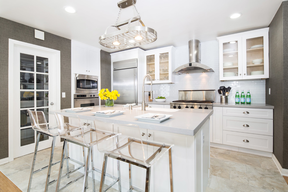 Medium sized traditional l-shaped kitchen/diner in Los Angeles with a belfast sink, shaker cabinets, engineered stone countertops, white splashback, stainless steel appliances, ceramic flooring, an island, white cabinets and metro tiled splashback.