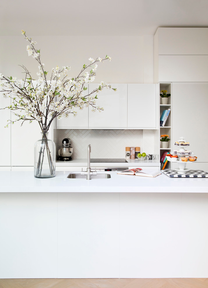 Photo of a medium sized contemporary galley open plan kitchen in London with a single-bowl sink, white cabinets, composite countertops, white splashback, mosaic tiled splashback, stainless steel appliances, light hardwood flooring and an island.