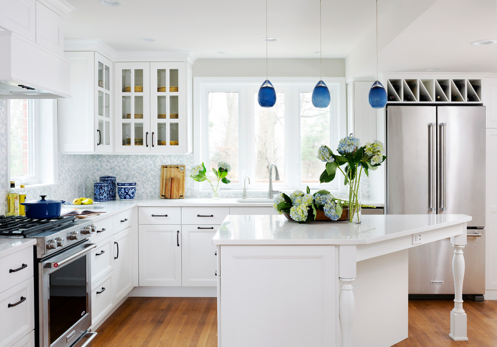 Klassisk inredning av ett mellanstort, avskilt l-kök, med en undermonterad diskho, luckor med glaspanel, vita skåp, bänkskiva i kvarts, blått stänkskydd, stänkskydd i mosaik, rostfria vitvaror, ljust trägolv, en köksö och beiget golv