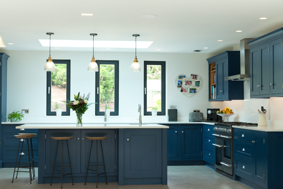 Classic l-shaped kitchen in Surrey with shaker cabinets, blue cabinets, white splashback, stainless steel appliances, an island, grey floors and white worktops.