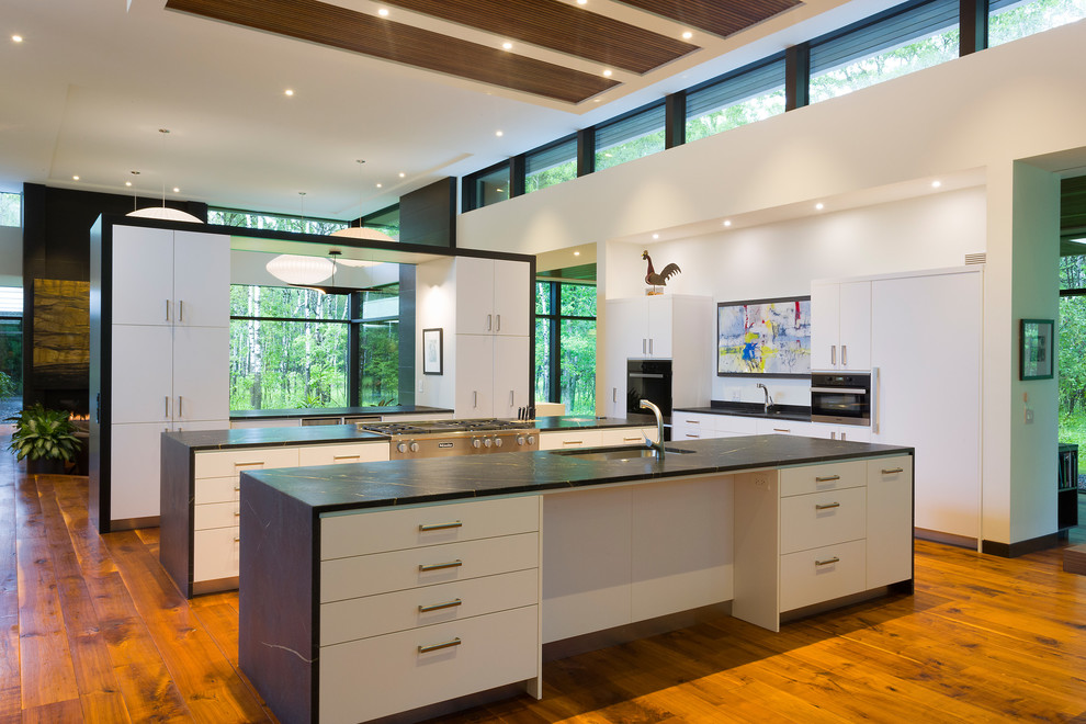 Inspiration for an expansive contemporary galley open plan kitchen in Other with multiple islands, a submerged sink, flat-panel cabinets, white cabinets, soapstone worktops, white splashback, stainless steel appliances, medium hardwood flooring, brown floors and black worktops.