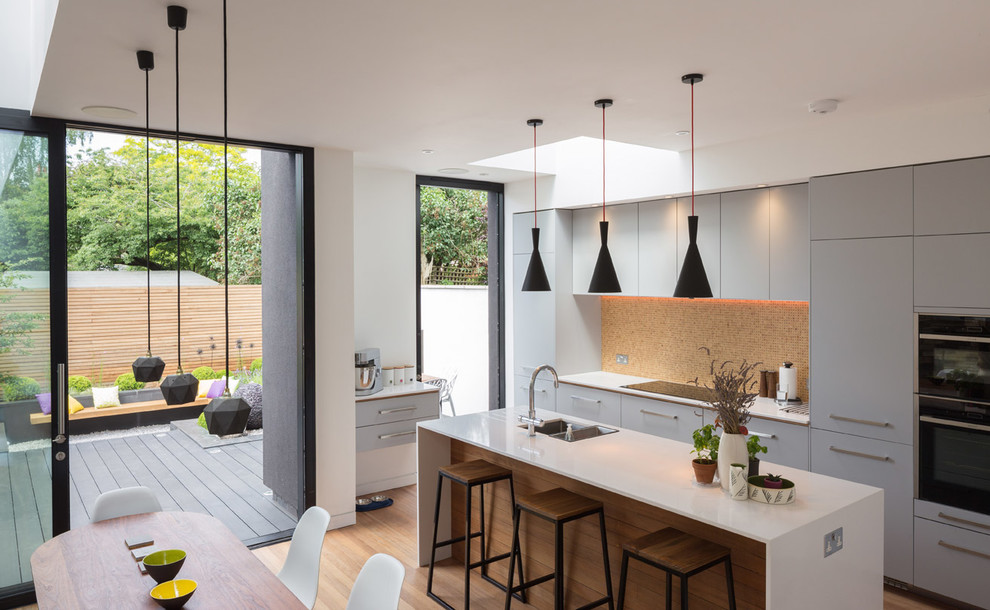 Photo of a small contemporary single-wall open plan kitchen in Other with a built-in sink, flat-panel cabinets, grey cabinets, multi-coloured splashback, mosaic tiled splashback, stainless steel appliances, medium hardwood flooring, an island and white worktops.
