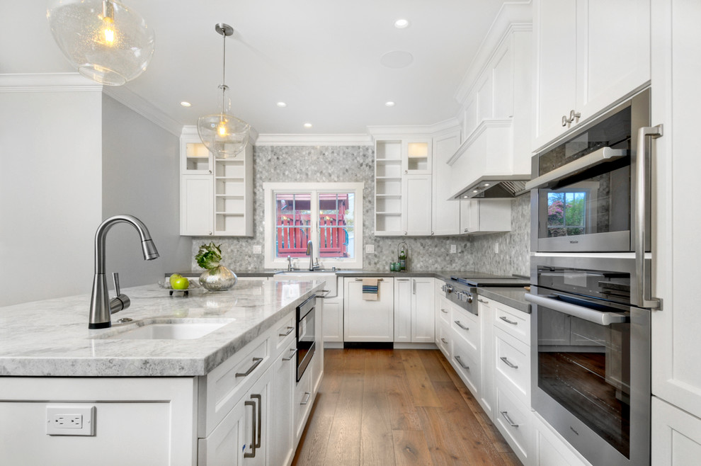 Kitchen - mid-sized transitional l-shaped dark wood floor and brown floor kitchen idea in San Francisco with a farmhouse sink, recessed-panel cabinets, white cabinets, solid surface countertops, gray backsplash, marble backsplash, paneled appliances, an island and gray countertops