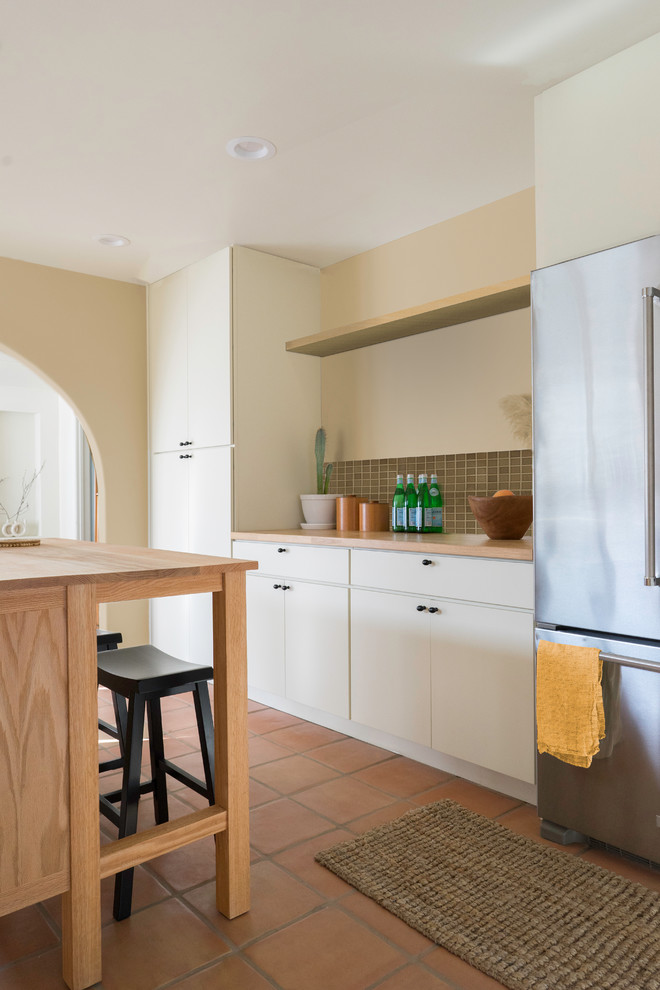 Foto på ett funkis brun linjärt kök och matrum, med släta luckor, vita skåp, träbänkskiva, grönt stänkskydd, stänkskydd i mosaik, rostfria vitvaror, klinkergolv i terrakotta, en köksö och brunt golv