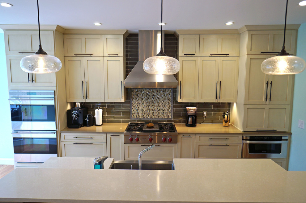 Photo of a medium sized traditional galley kitchen/diner in Boston with a belfast sink, shaker cabinets, beige cabinets, engineered stone countertops, glass tiled splashback, stainless steel appliances, medium hardwood flooring, an island and brown splashback.