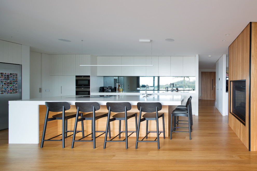 Offene Moderne Küche in L-Form mit flächenbündigen Schrankfronten, Rückwand aus Spiegelfliesen, schwarzen Elektrogeräten, braunem Holzboden, Kücheninsel und weißer Arbeitsplatte in Sydney