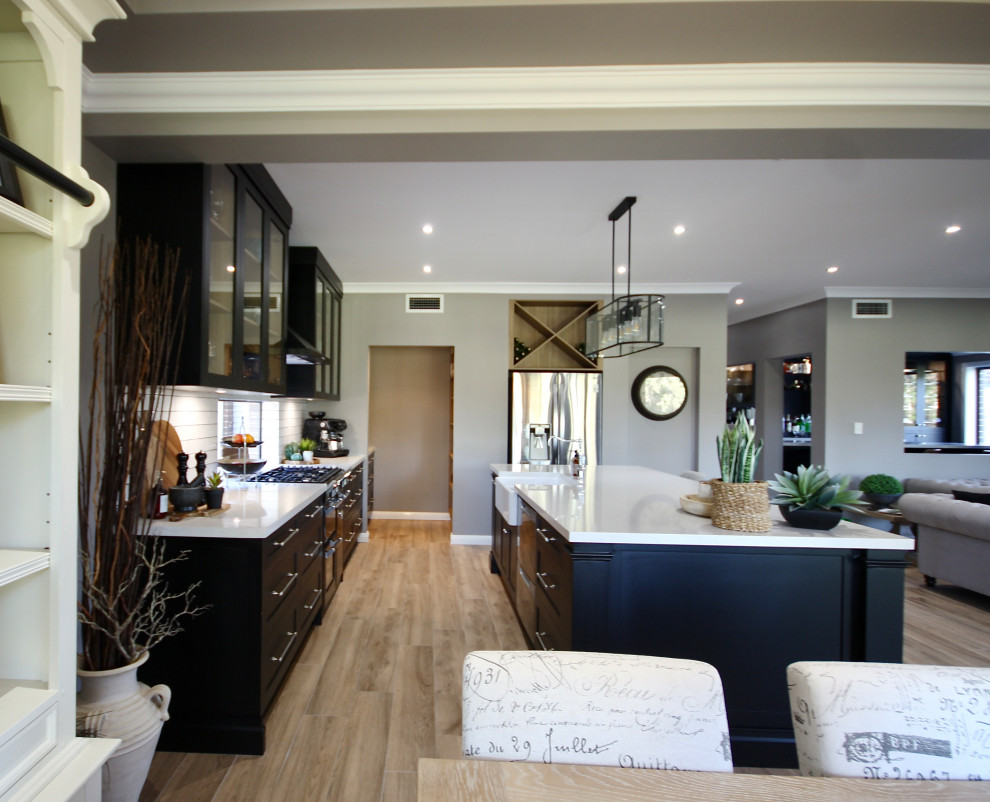 Design ideas for a large traditional single-wall open plan kitchen in Sydney with a belfast sink, shaker cabinets, black cabinets, engineered stone countertops, white splashback, metro tiled splashback, black appliances, light hardwood flooring, an island, brown floors, white worktops and a coffered ceiling.
