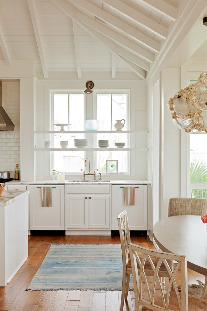 Imagen de cocina costera con armarios abiertos, encimera de mármol, salpicadero blanco, salpicadero de azulejos de piedra, electrodomésticos de acero inoxidable, suelo de madera en tonos medios y una isla