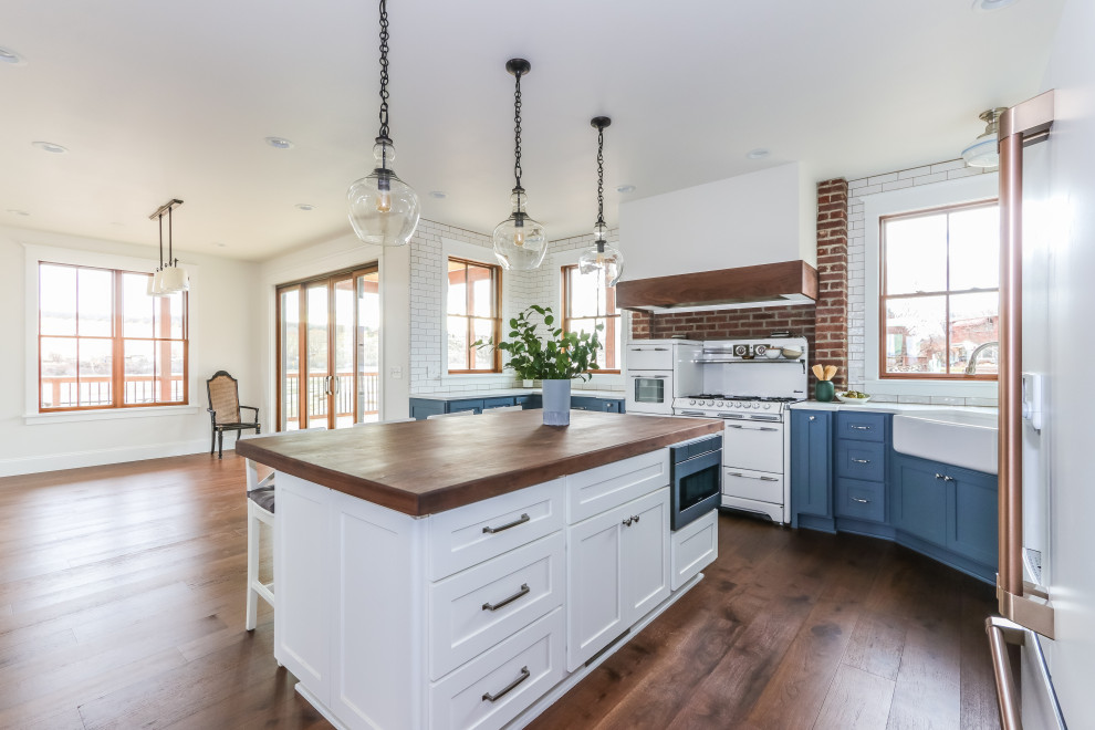 Inspiration for a large farmhouse u-shaped kitchen/diner in Seattle with a belfast sink, shaker cabinets, blue cabinets, engineered stone countertops, white splashback, metro tiled splashback, white appliances, medium hardwood flooring, an island, brown floors and white worktops.
