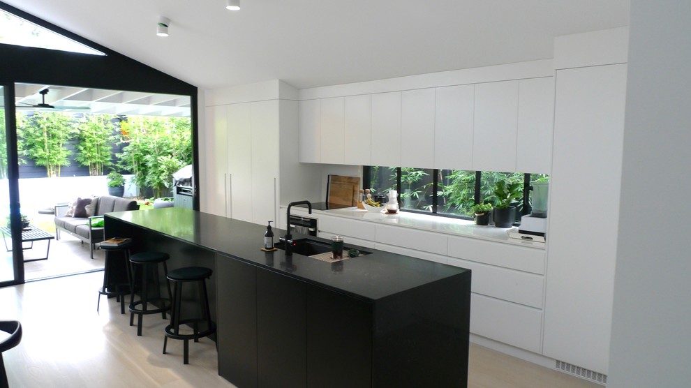 Design ideas for a medium sized modern galley open plan kitchen in Sydney with a submerged sink, flat-panel cabinets, black cabinets, composite countertops, white splashback, glass sheet splashback, black appliances, painted wood flooring, an island, white floors and white worktops.