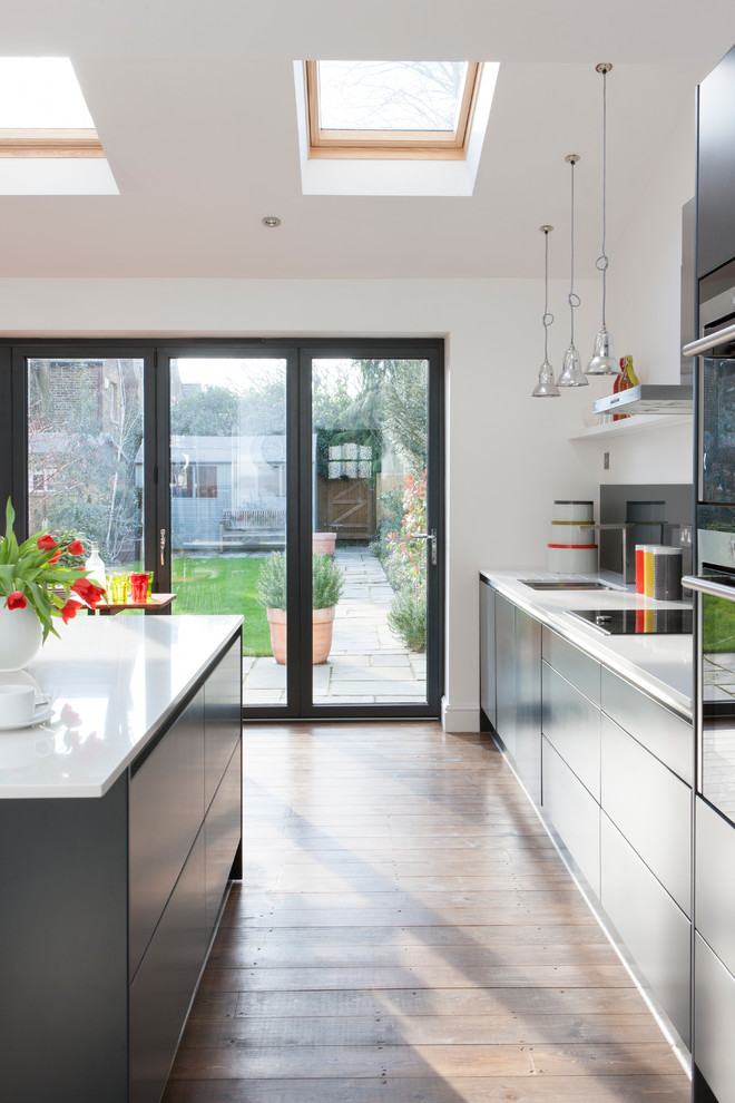 This is an example of a medium sized contemporary single-wall kitchen/diner in London with an integrated sink, flat-panel cabinets, grey cabinets, composite countertops, grey splashback, glass sheet splashback, integrated appliances, an island, brown floors, white worktops and feature lighting.
