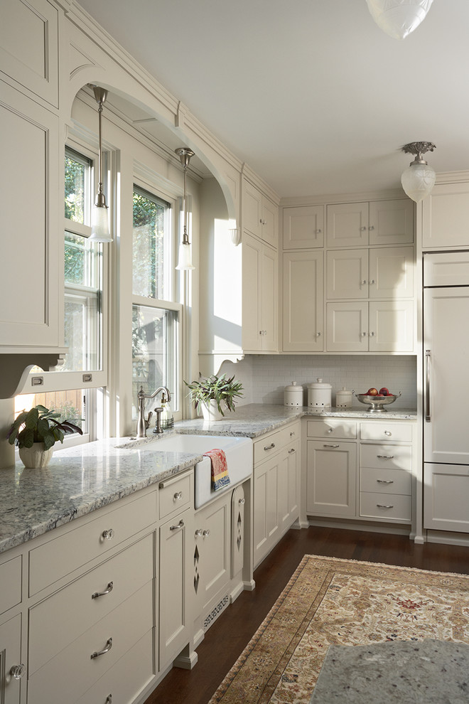 Example of a mid-sized ornate dark wood floor and brown floor kitchen design in Minneapolis with a farmhouse sink, recessed-panel cabinets, white cabinets, granite countertops, white backsplash, ceramic backsplash and paneled appliances