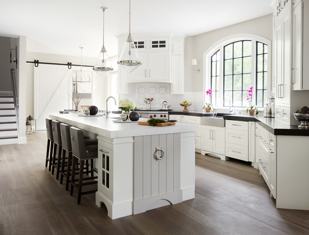 Photo of a large traditional u-shaped kitchen/diner in Chicago with a belfast sink, shaker cabinets, white cabinets, white splashback, metro tiled splashback, stainless steel appliances, medium hardwood flooring, an island and composite countertops.