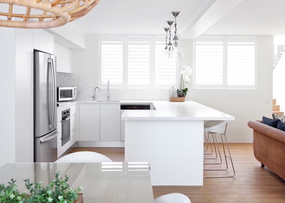 Bild på ett funkis vit vitt kök, med släta luckor, vita skåp, grått stänkskydd, rostfria vitvaror, mellanmörkt trägolv, en halv köksö och brunt golv