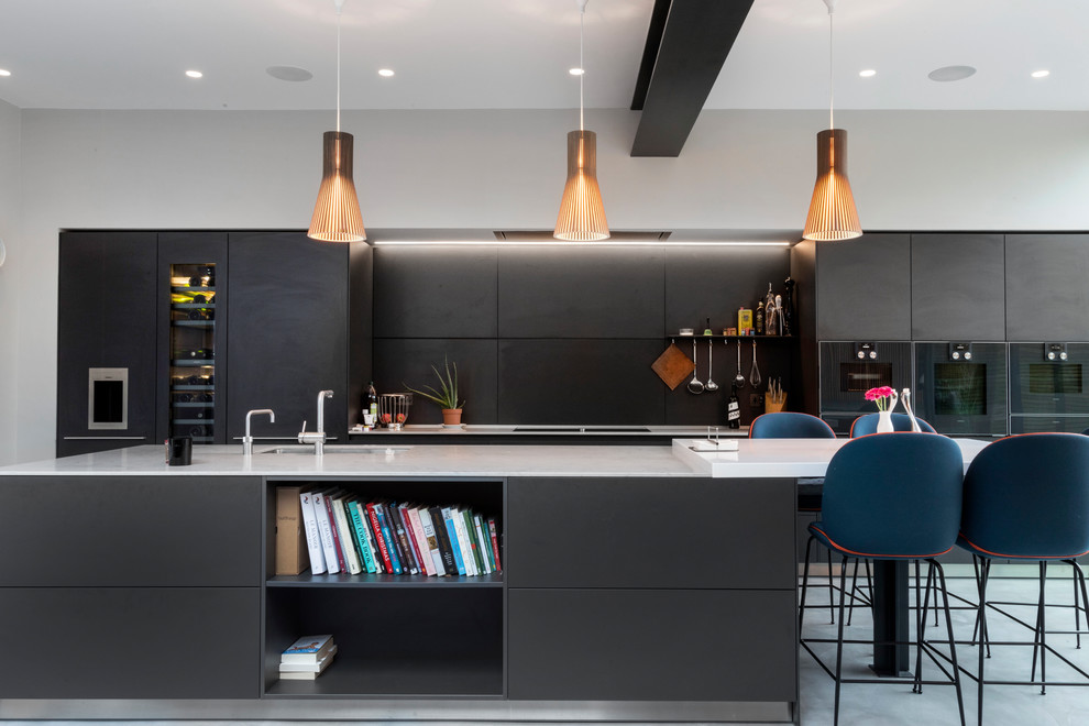 Eat-in kitchen - contemporary concrete floor and gray floor eat-in kitchen idea in London with an undermount sink, flat-panel cabinets, black cabinets, black backsplash, paneled appliances, an island and white countertops