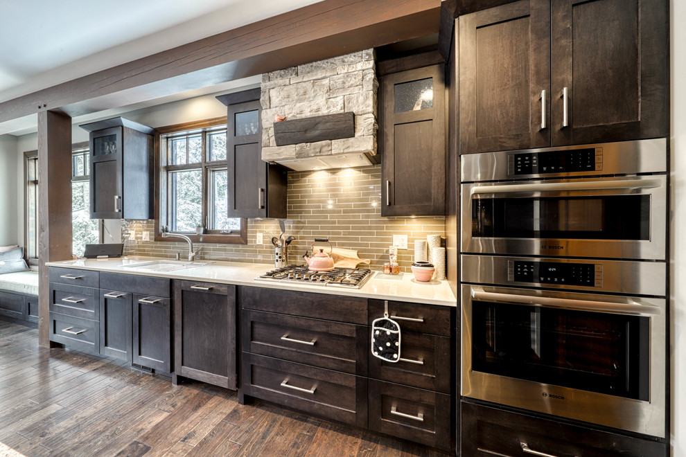 Eat-in kitchen - large rustic l-shaped dark wood floor and brown floor eat-in kitchen idea in Vancouver with a drop-in sink, recessed-panel cabinets, dark wood cabinets, brown backsplash, stainless steel appliances, an island and white countertops