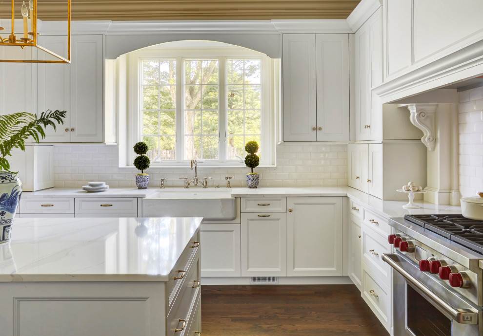 Bild på ett mycket stort vintage vit vitt kök, med en rustik diskho, luckor med upphöjd panel, vita skåp, bänkskiva i kvarts, vitt stänkskydd, stänkskydd i mosaik, integrerade vitvaror, mellanmörkt trägolv, en köksö och brunt golv