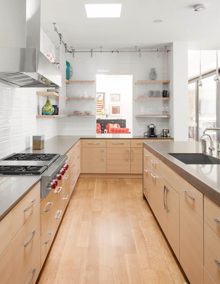 Cette image montre une cuisine design en bois clair avec un placard à porte plane, îlot, un évier encastré, une crédence blanche, un électroménager en acier inoxydable et parquet clair.