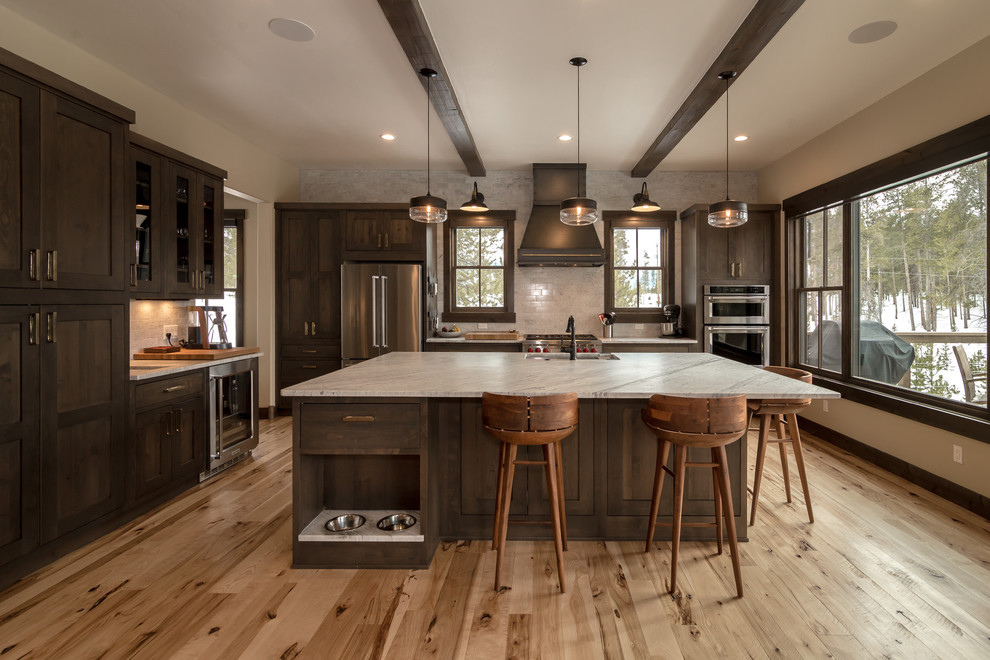 This is an example of a large rustic l-shaped open plan kitchen in Denver with a submerged sink, shaker cabinets, dark wood cabinets, engineered stone countertops, beige splashback, metro tiled splashback, stainless steel appliances, light hardwood flooring, an island, beige floors and white worktops.