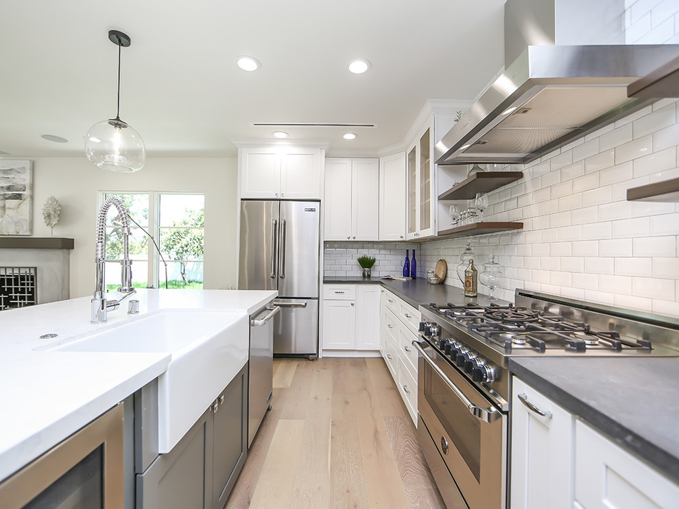Example of a mid-sized beach style l-shaped light wood floor open concept kitchen design in Los Angeles with a farmhouse sink, shaker cabinets, white cabinets, marble countertops, white backsplash, subway tile backsplash, stainless steel appliances and an island