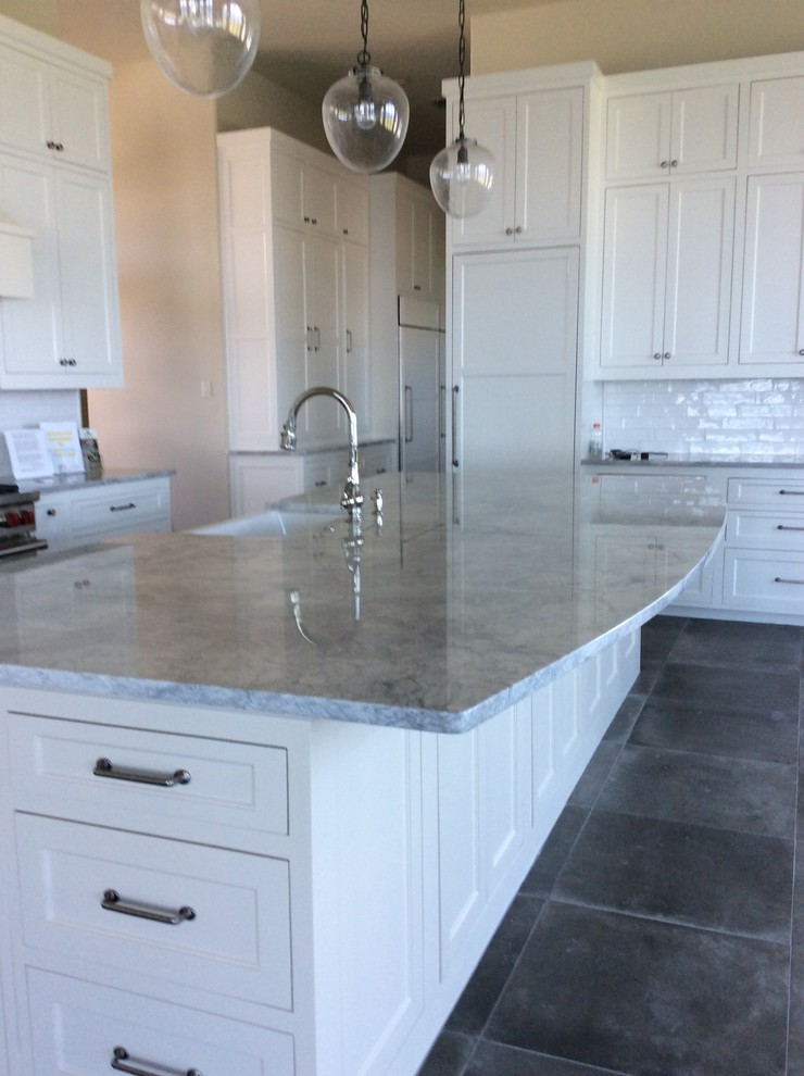 Example of a large minimalist u-shaped travertine floor enclosed kitchen design in Other with a farmhouse sink, recessed-panel cabinets, gray cabinets, quartzite countertops, white backsplash, subway tile backsplash, stainless steel appliances and an island