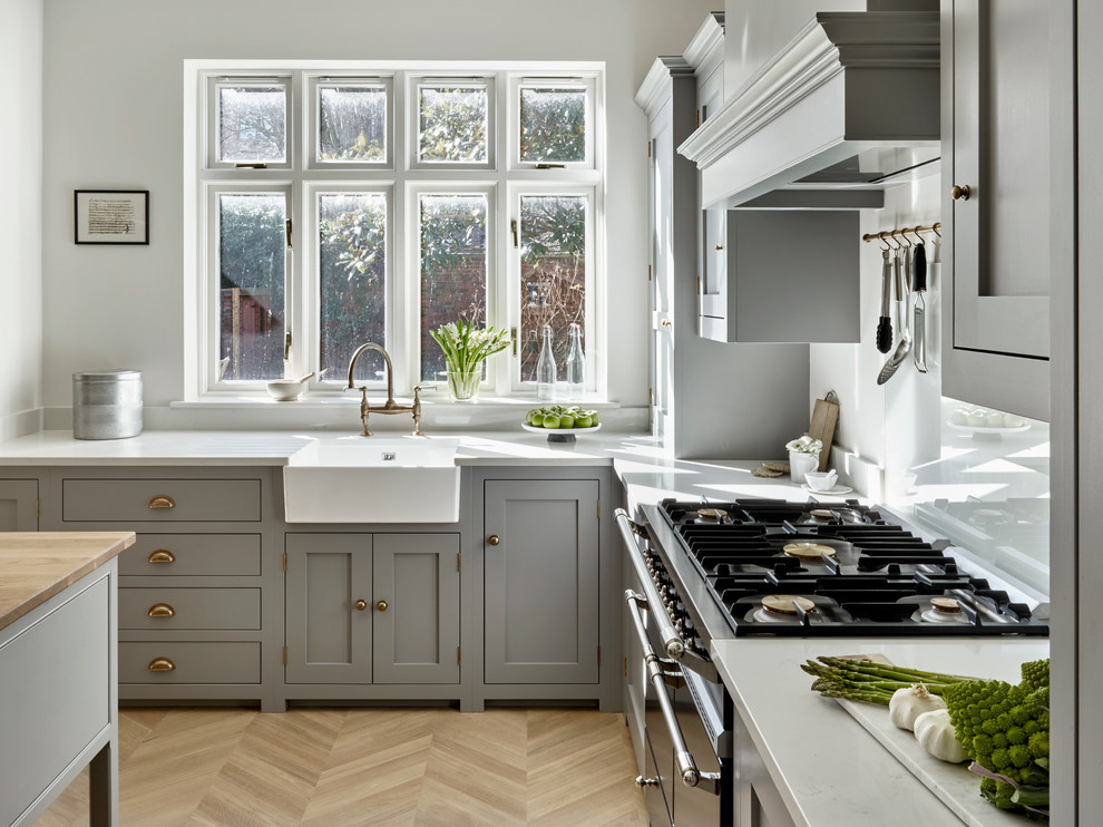 Photo of a classic l-shaped kitchen/diner in London with a belfast sink, shaker cabinets, grey cabinets, light hardwood flooring, an island and white worktops.