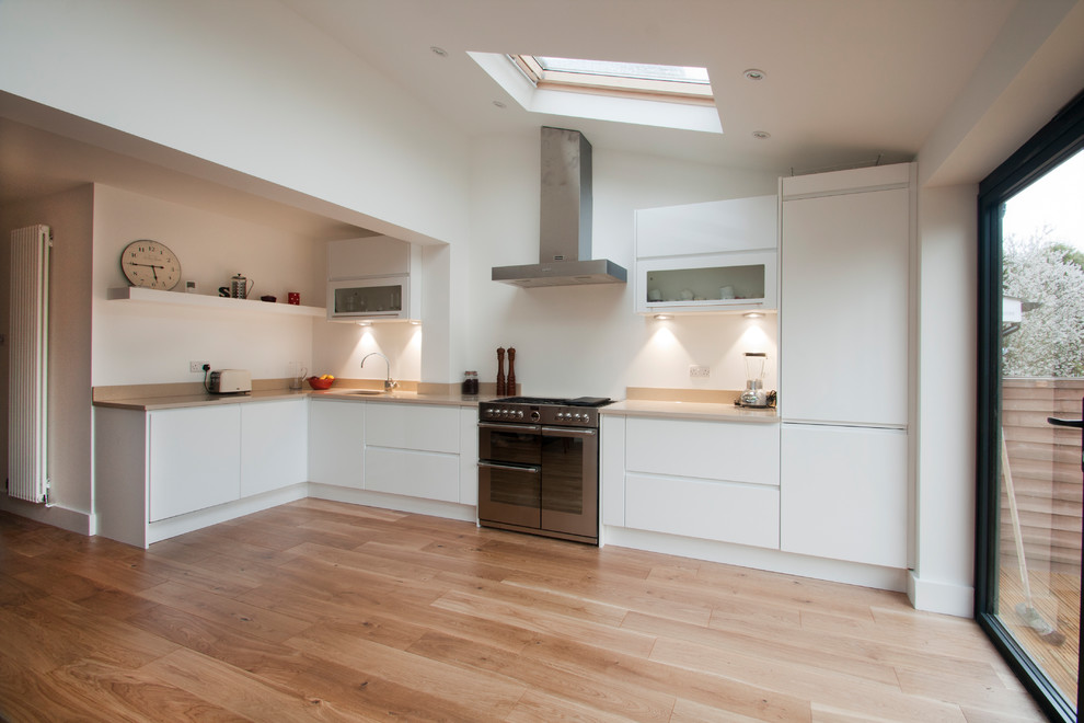 This is an example of a small contemporary l-shaped open plan kitchen in Surrey with a built-in sink, flat-panel cabinets, white cabinets, granite worktops, stainless steel appliances, light hardwood flooring, no island and beige worktops.