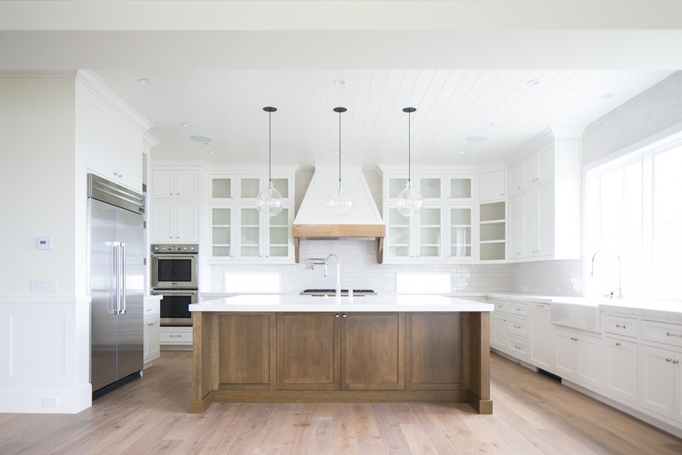 Example of a large transitional u-shaped light wood floor and brown floor open concept kitchen design in Orange County with a farmhouse sink, shaker cabinets, white cabinets, marble countertops, white backsplash, subway tile backsplash, stainless steel appliances and an island