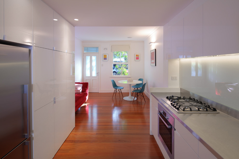 Modern inredning av ett kök, med en integrerad diskho, släta luckor, vita skåp, bänkskiva i rostfritt stål, vitt stänkskydd, glaspanel som stänkskydd och rostfria vitvaror