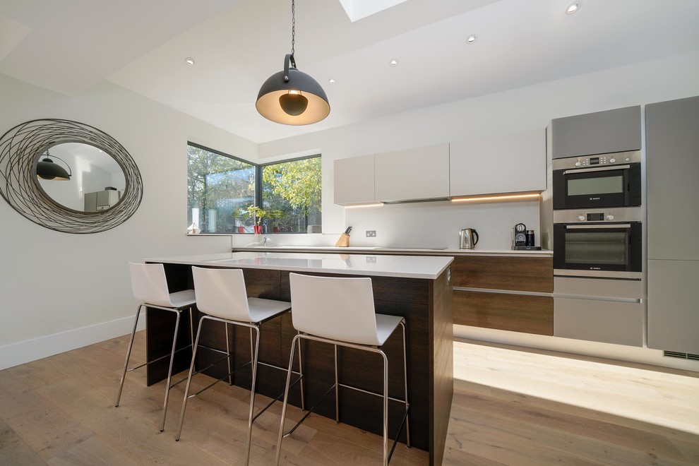 Photo of a medium sized contemporary single-wall open plan kitchen in London with a built-in sink, flat-panel cabinets, dark wood cabinets, quartz worktops, white splashback, glass sheet splashback, stainless steel appliances, light hardwood flooring, an island and white worktops.