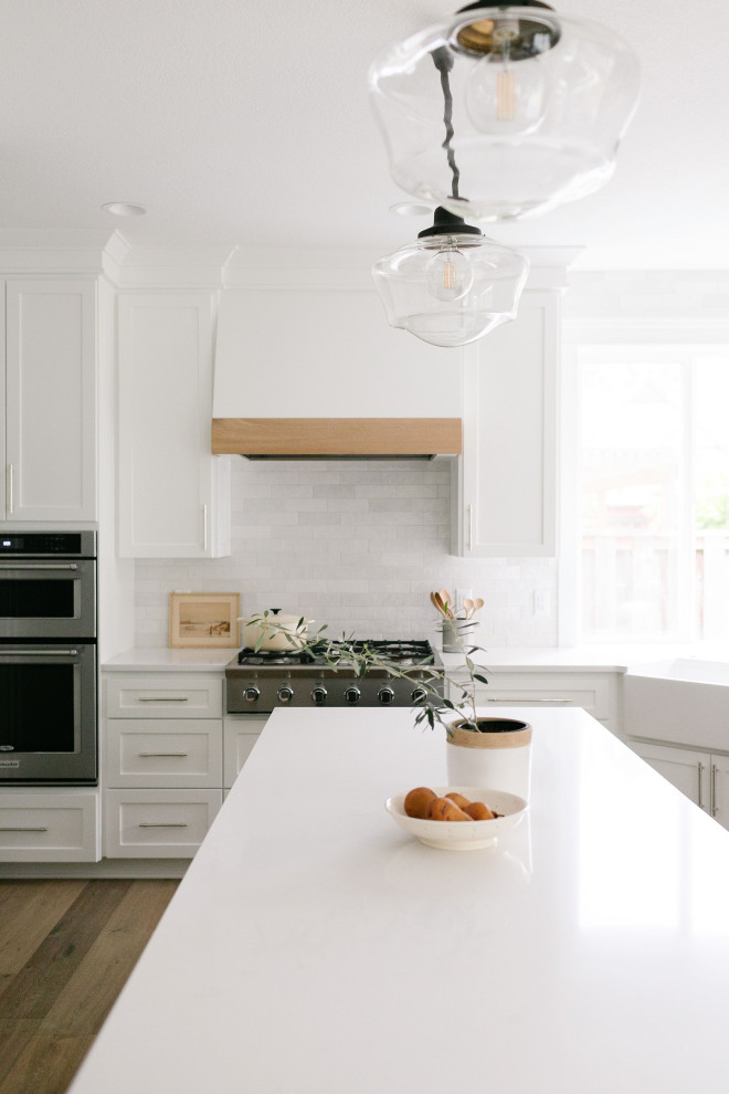 Bild på ett stort amerikanskt vit vitt kök, med en rustik diskho, skåp i shakerstil, vita skåp, bänkskiva i kvartsit, vitt stänkskydd, stänkskydd i keramik, rostfria vitvaror, ljust trägolv, en köksö och brunt golv