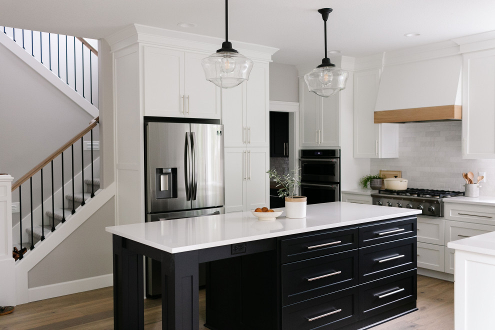 Large arts and crafts u-shaped light wood floor and brown floor open concept kitchen photo in Portland with a farmhouse sink, shaker cabinets, white cabinets, quartzite countertops, white backsplash, ceramic backsplash, stainless steel appliances, an island and white countertops