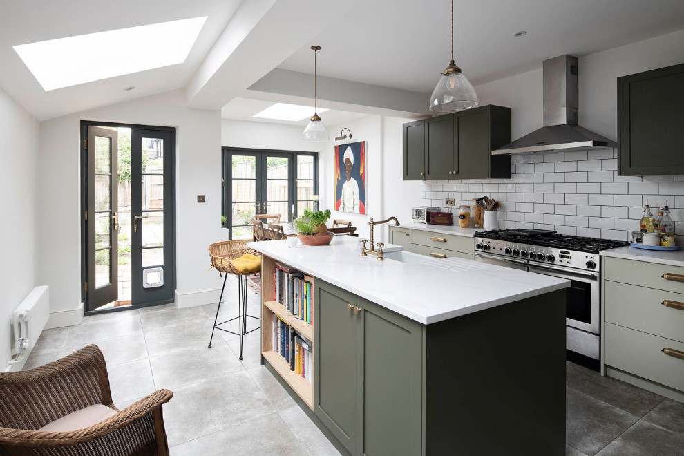 This is an example of a traditional galley kitchen/diner in London with a belfast sink, shaker cabinets, green cabinets, white splashback, metro tiled splashback, stainless steel appliances, an island, grey floors and white worktops.