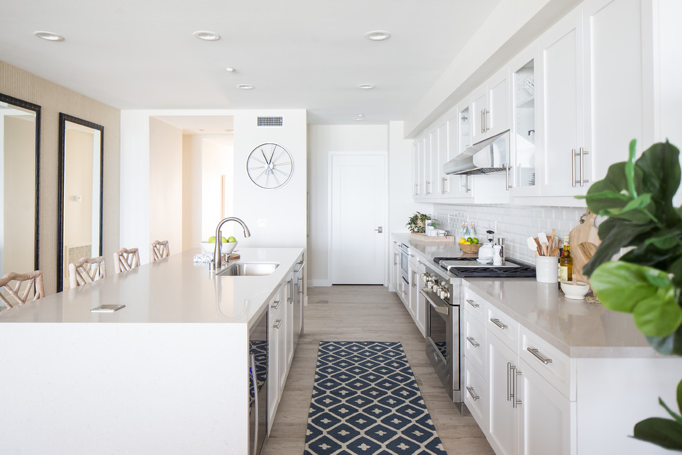 Modelo de cocina lineal costera con puertas de armario blancas, salpicadero blanco, salpicadero de azulejos tipo metro, electrodomésticos de acero inoxidable y una isla