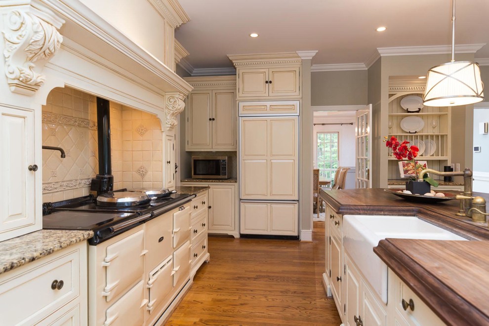 Large elegant l-shaped medium tone wood floor and brown floor eat-in kitchen photo in Raleigh with a farmhouse sink, raised-panel cabinets, beige cabinets, wood countertops, beige backsplash, porcelain backsplash, paneled appliances, an island and brown countertops