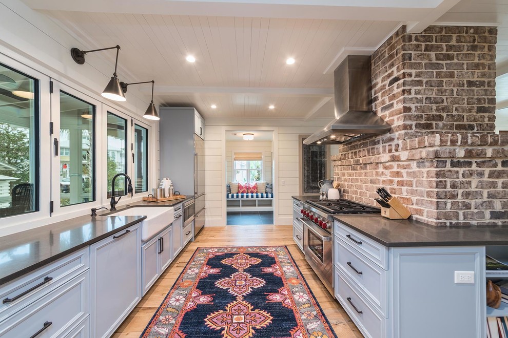 Photo of a large beach style l-shaped kitchen in Atlanta with a belfast sink, recessed-panel cabinets, grey cabinets, composite countertops, stainless steel appliances, light hardwood flooring, an island, brown floors and grey worktops.