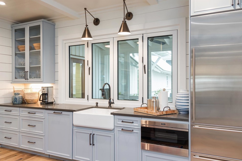 This is an example of a large beach style l-shaped kitchen in Atlanta with a belfast sink, recessed-panel cabinets, grey cabinets, composite countertops, stainless steel appliances, light hardwood flooring, an island, brown floors and grey worktops.