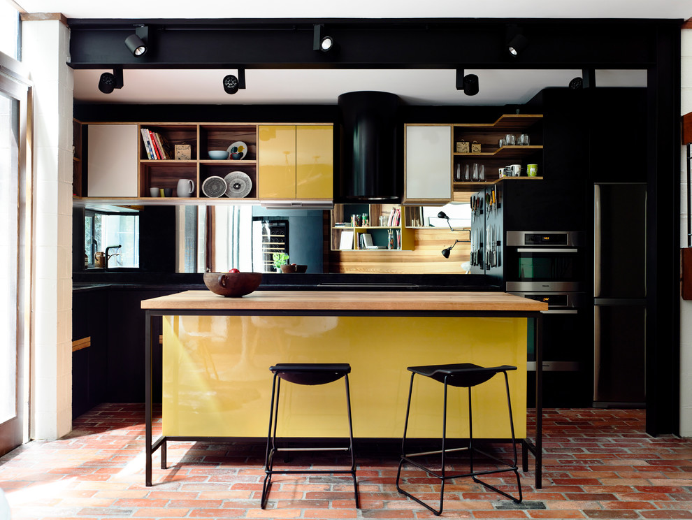 This is an example of a medium sized eclectic l-shaped kitchen/diner in Melbourne with flat-panel cabinets, yellow cabinets, an island, a submerged sink, wood worktops, black splashback, mirror splashback, brick flooring and stainless steel appliances.