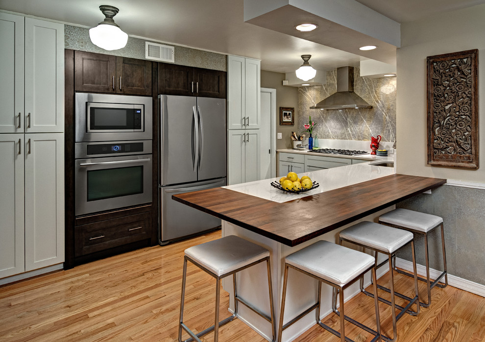Inspiration for a mid-sized transitional u-shaped medium tone wood floor kitchen remodel in Minneapolis with an undermount sink, shaker cabinets, white cabinets, wood countertops, beige backsplash, stone slab backsplash, stainless steel appliances and a peninsula