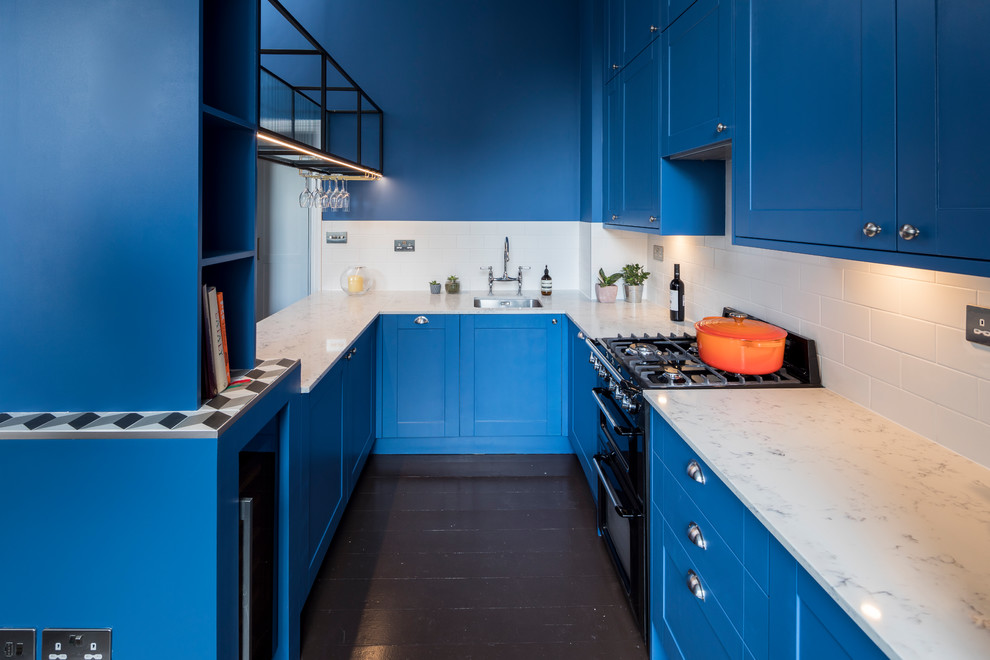 Inspiration for a traditional u-shaped kitchen in London with a submerged sink, shaker cabinets, blue cabinets, white splashback, metro tiled splashback, black appliances, a breakfast bar, brown floors and white worktops.
